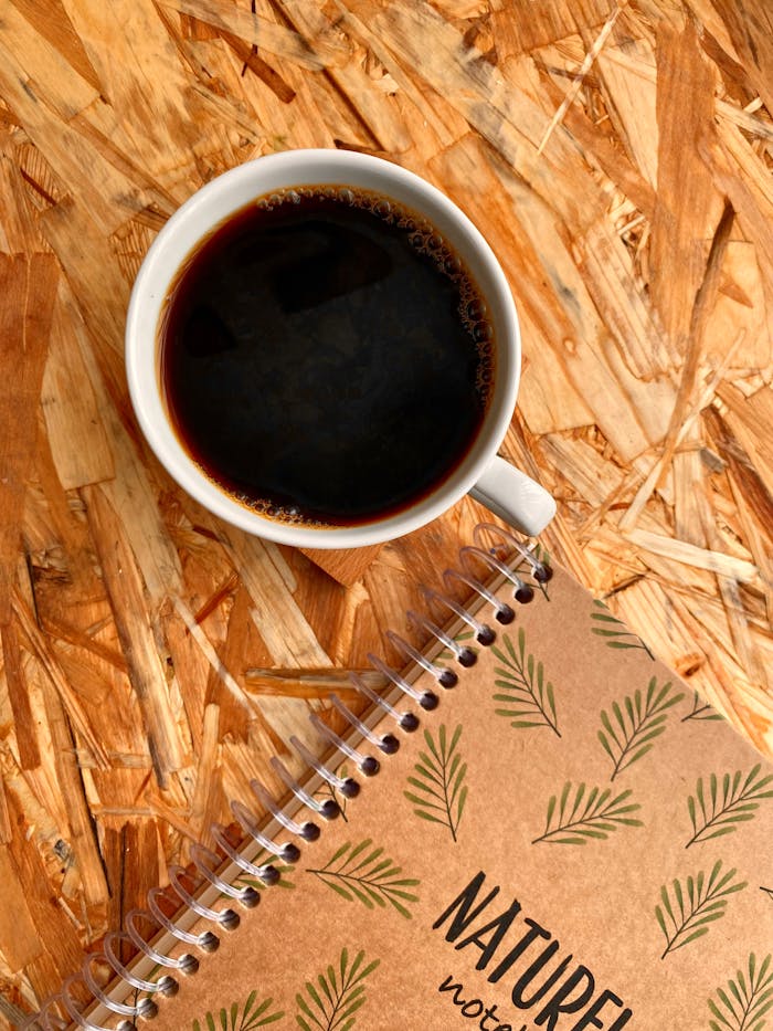 A coffee cup and spiral notebook placed on a wooden textured surface.