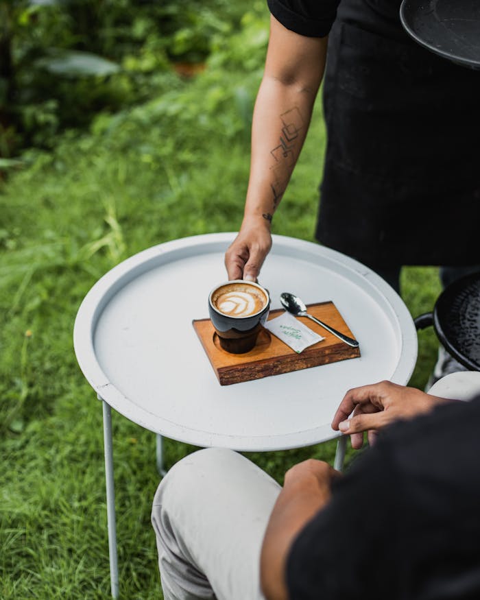 A barista serving a cup of latte coffee with art on a small round table outdoors.